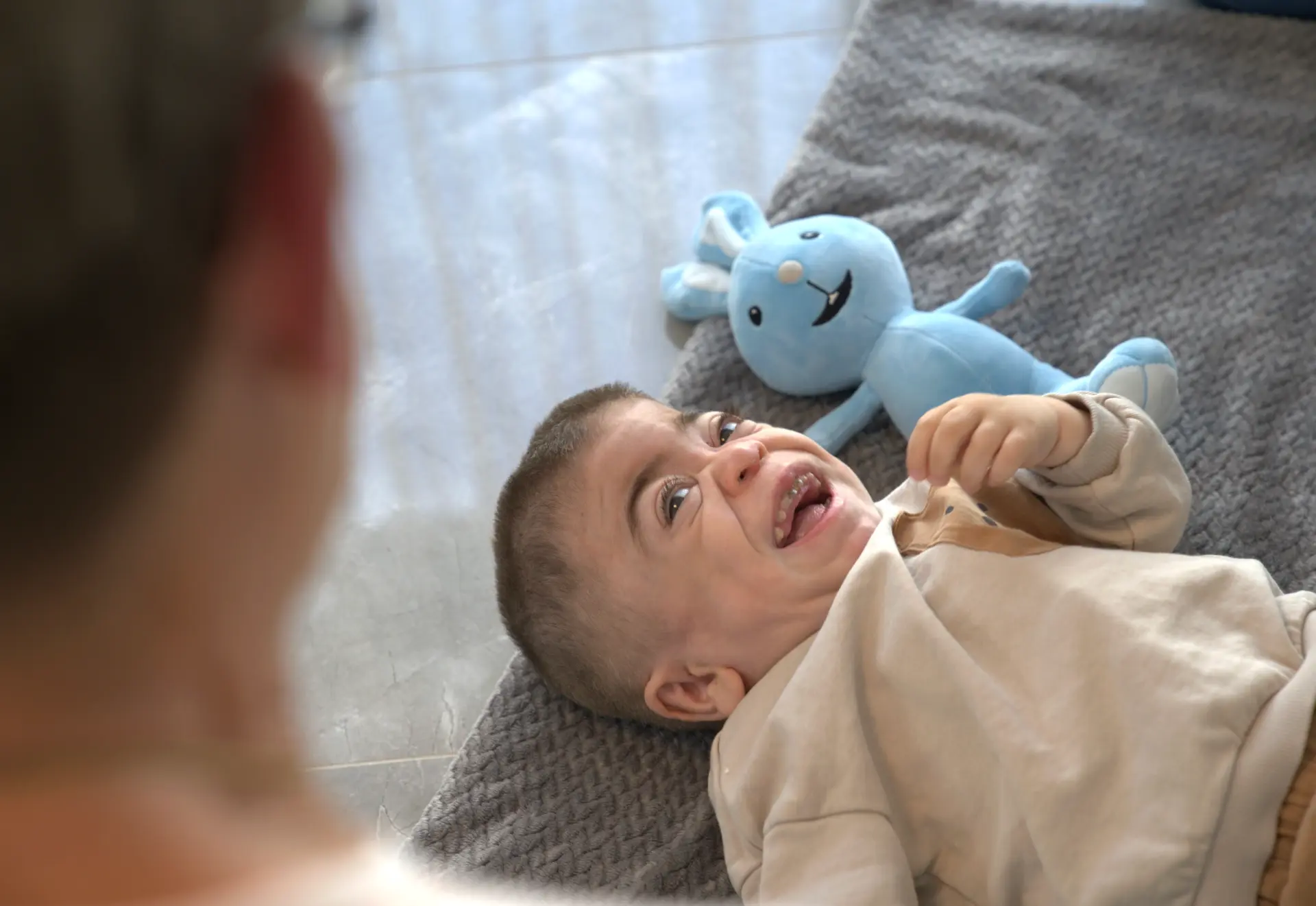 Lachendes Kind liegt mit blauem Kuscheltier auf einem Kissen zuhause